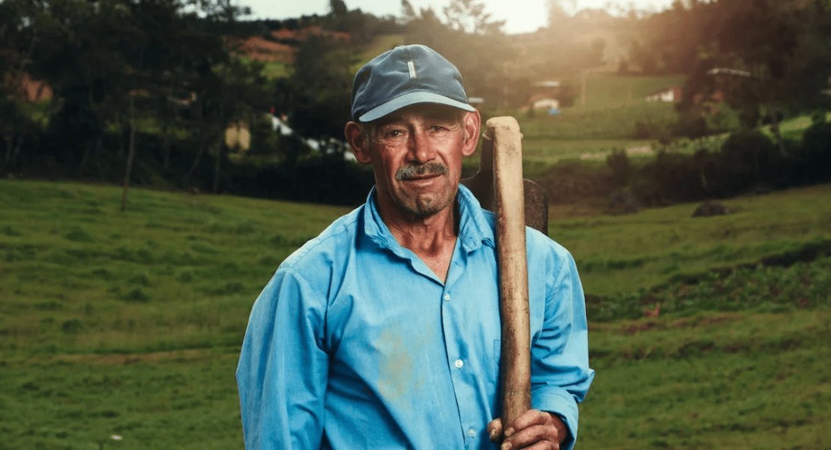 Trabalhador do campo de boné e camisa azul com uma ferramenta apoiada no ombro, num pasto verde com árvores ao fundo