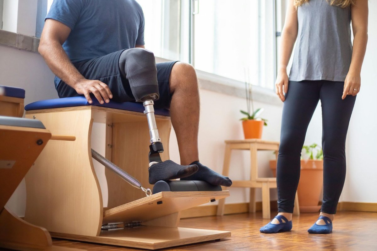 Homem com prótese na perna sentado em um aparelho de fisioterapia, ao lado de uma profissional