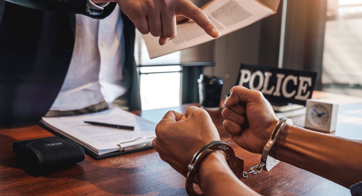 Mãos algemadas sobre uma mesa enquanto um homem de terno aponta para um documento