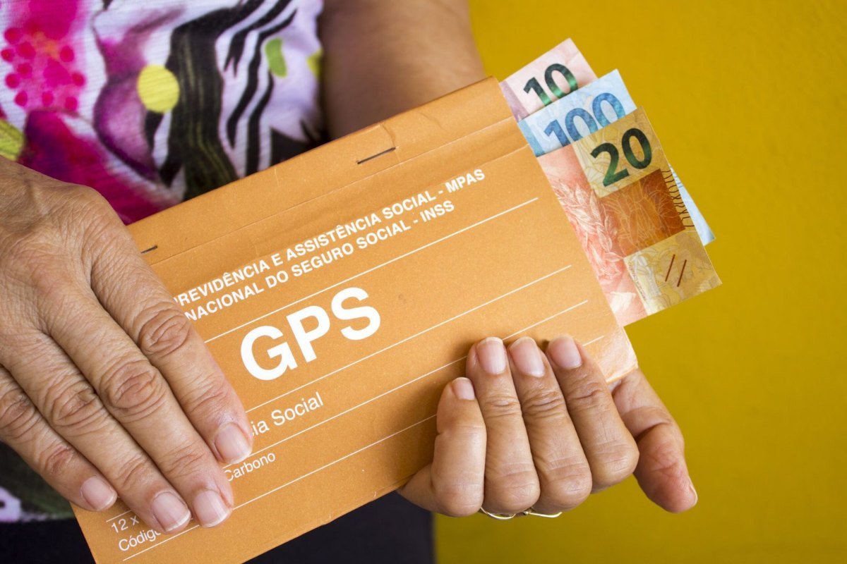 Mãos segurando um carnê de GPS do INSS e cédulas de real